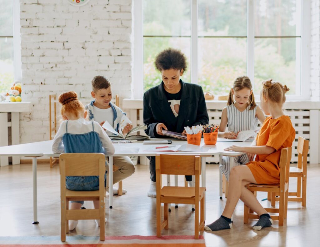 Children learning with a teacher around a table in a bright classroom setting.